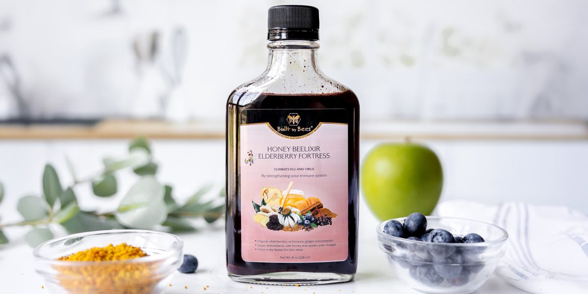 Bottle of elderberry honey elixir on countertop with bowl of bee pollen, fresh blueberries and a green apple