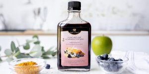 Bottle of elderberry honey elixir on countertop with bowl of bee pollen, fresh blueberries and a green apple