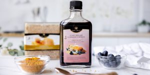 Bottle of elderberry honey elixir on marble counter with bee pollen in a bowl, fresh blueberries, and honeycomb box blurred in background