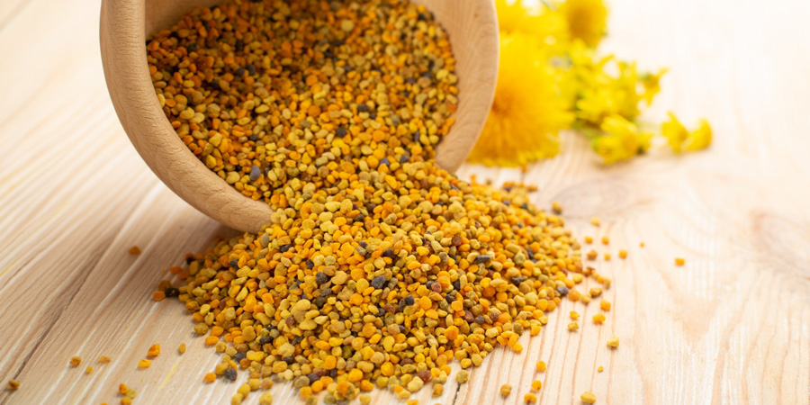 Bee pollen granules spilling from a wooden bowl onto a light wooden surface with yellow wildflowers blurred in background