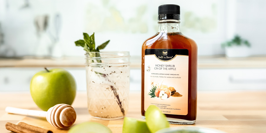 Bottle of apple honey shrub on wooden counter with green apples, honey dipper, and a mason jar cocktail garnished with mint