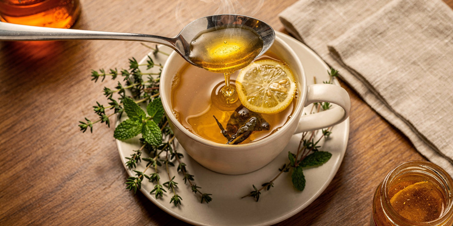 Steaming cup of lemon ginger tea with honey drizzling from a spoon, fresh mint and thyme sprigs on saucer