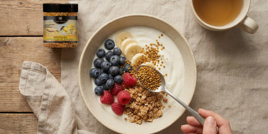 Hand sprinkling bee pollen over yogurt bowl with granola, blueberries, raspberries and banana slices beside a Built by Bees bee pollen jar and cup of tea