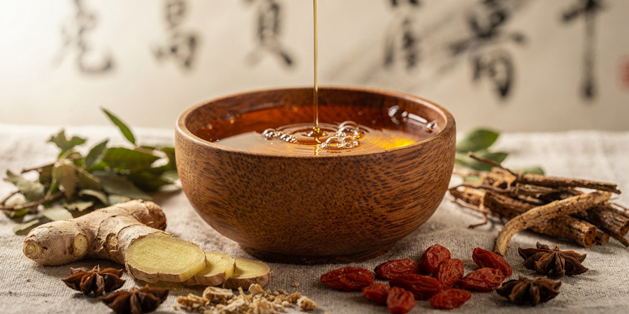 Honey pouring into wooden bowl surrounded by sliced ginger, goji berries, star anise and herbs—natural remedy and herbal honey close-up