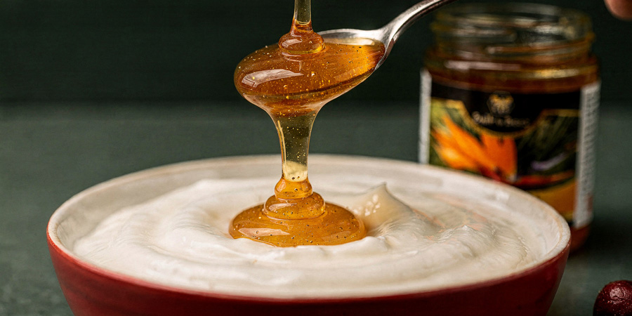 Golden honey drizzling from a spoon into creamy yogurt in a red bowl with a jar in background
