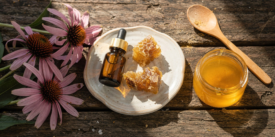 Jar of golden honey, honeycomb pieces and propolis bottle on a ceramic plate beside pink echinacea flowers and wooden spoon on rustic wood