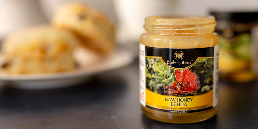 Jar of raw Lehua honey with bright floral label on a counter, blurred scones in background