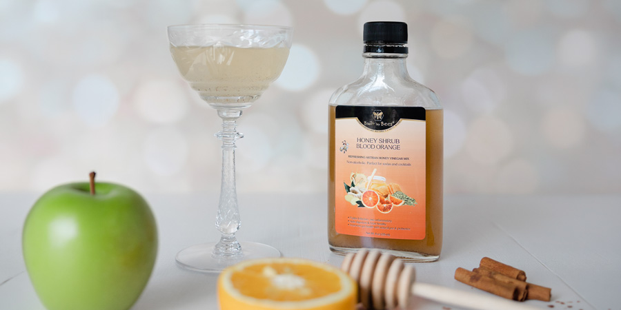 Bottle of blood orange honey shrub beside a cocktail glass, green apple, sliced orange, honey dipper and cinnamon sticks on white table