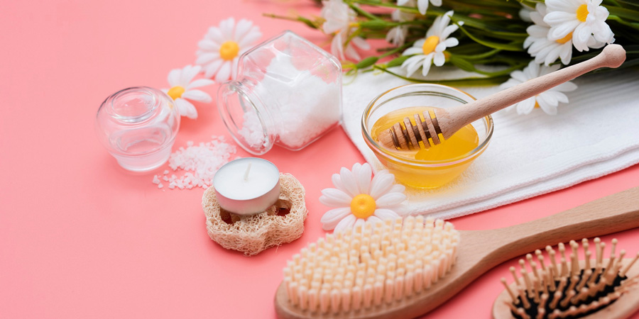 Spa essentials on pink background: honey in glass bowl with wooden dipper, sea salt jar, loofah, hairbrushes and daisies for natural skincare routine.