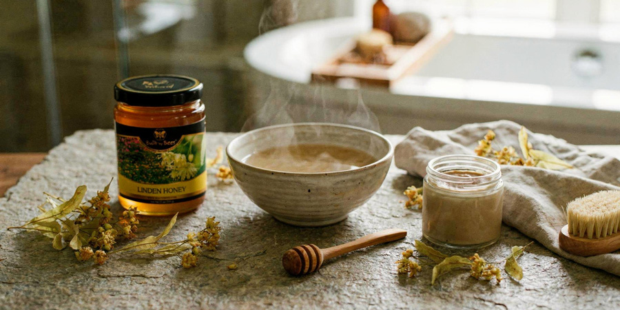 Jar of linden honey, steaming bowl of tea, honey dipper and natural skincare jar with dried linden blossoms on a rustic surface