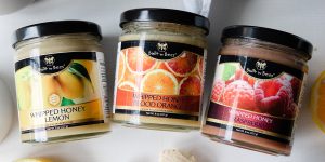 Three jars of Built By Bees whipped honey (lemon, blood orange, raspberry) arranged on a light surface with citrus and berry accents, clear product labels visible, bright natural lighting, clean lifestyle product shot.