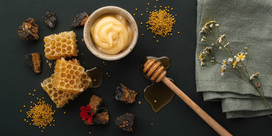 Flatlay of honey products: honeycomb pieces, whipped honey in a bowl, honey dipper with drips, bee pollen granules and wildflowers on dark backdrop